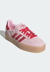 Růžová semišová teniska s červenými pruhy, šněrováním vpředu a růžovým podš�ívkou. Má průsvitnou gumovou podrážku a nápis "Samba" na boku.