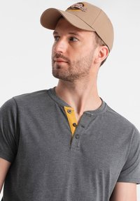 Camiseta henley gris de manga corta con una tapeta de botones amarillos. Gorra beige con un diseño bordado en la parte frontal. Textura de tela suave.