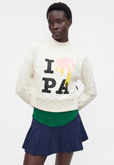 Γυναίκα που φοράει λευκό φούτερ με το κείμενο «I PA» και γραφικό σχεδιασμό με ροζ-κίτρινο φοίνικα, συνδυασμένο με πράσινο τοπ και σκούρα μπλε πλισέ φούστα.