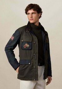 Muškarac u tamnozelenoj vojnog stilu jakni s našivenim oznakama, crnom rolkom i bež hlačama, gleda ulijevo pred jednostavnom pozadinom.