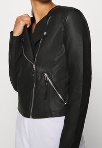 Giacca da motociclista nera in pelle con un design aderente, zip asimmetrica e tasche con zip. Presenta un materiale testurizzato e dettagli in metallo argentato.