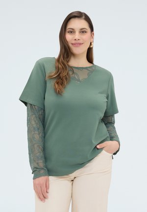 Lächelnde Frau mit langen Haaren, die goldene Creolen, ein grünes Oberteil mit transparenten Blumenärmeln und hellbeige Hose trägt, posiert mit einer Hand in der Hosentasche.