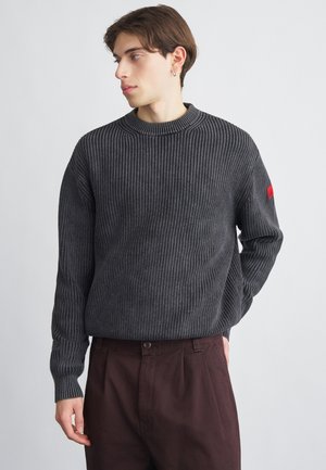 Tumehall triibuline kampsun ümar kaelusega ja pikkade varrukatega. Vasakul varrukal on punane aktsent. Ühendas pruuni pükstega.