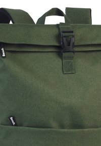 Sac à dos en tissu vert avec une fermeture enroulable, boucle en plastique noir et une petite étiquette de marque. Matériau texturé et bretelles ajustables.
