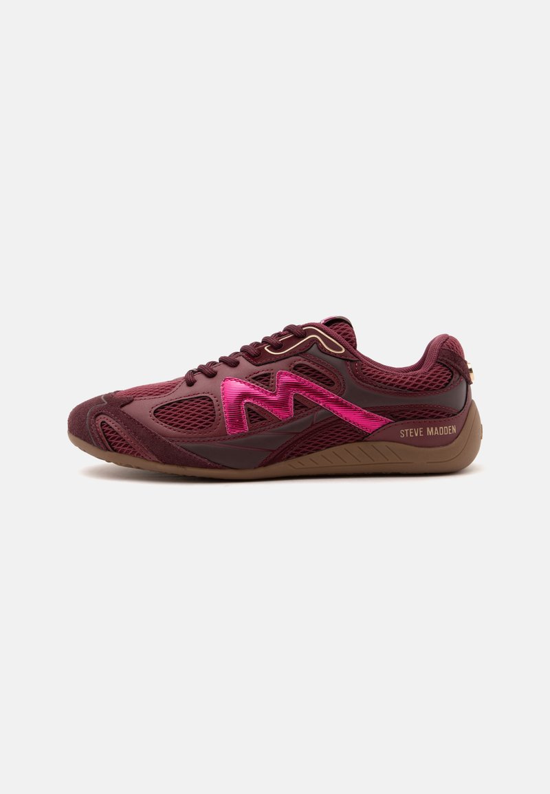 Une chaussure de sport marron foncé avec des panneaux en mesh, logo rose "M" sur le côté et semelle marron clair marquée "Steve Madden" au talon.
