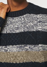 Pletený svetr v námořnické modři, krémové a béžové barvě s texturovanými kabelovými vzory a žebrovaným kulatým výstřihem.