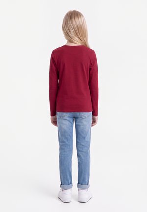 Enfant aux longs cheveux blonds, de dos, portant un haut rouge à manches longues, un jean bleu clair et des baskets blanches sur fond blanc.