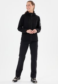 Sort softshell-jakke med lynlås foran, høj krave og sidelommer; parret med sorte bukser og grå vandresko.