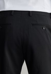 Vue arrière d’un pantalon noir formel avec deux poches arrière boutonnées et des passants de ceinture, porté avec une chemise blanche rentrée.