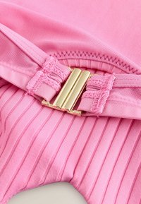 Lyserødt ribbet stof med en glat tekstur, der har en lukning med en tre-stangs spænde i guldton og dekorative syningsdetaljer.