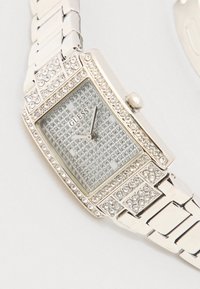 Ασημένιο ορθογώνιο ρολόι με διακοσμήσεις από στρας, μεταλλικό μπρασελέ και υφή στο καντράν που διαθέτει μικρά τετράγωνα σημάδια και το όνομα της μάρκας "GUESS".