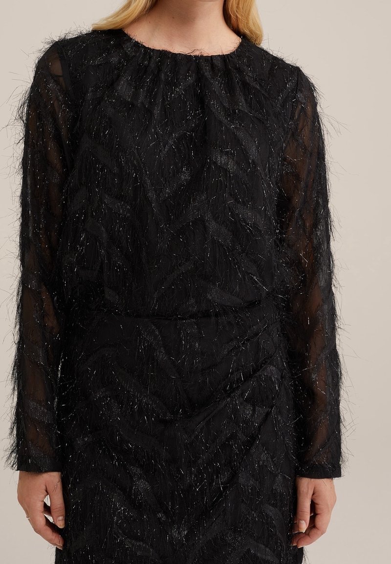 Haut noir à manches longues avec manches transparentes et détails en fil texturé et brillant porté par une personne à la peau claire et aux cheveux blonds.