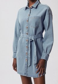 Vestido camisero de mezclilla en azul claro, con un frente abotonado, un bolsillo en el pecho, mangas largas y un cinturón a juego atado en la cintura.