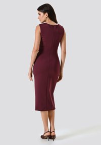 Burgunderfarbenes ärmelloses Kleid mit tailliertem Design, knielangem Schnitt und Reißverschlussdetails im Rücken. Ein Paar braune High Heels rundet den Look ab.