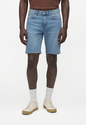 Hombre con pantalones cortos vaqueros azul claro, calcetines blancos hasta la mitad de la pantorrilla y zapatillas blancas con suelas de goma, de pie frente a un fondo liso.