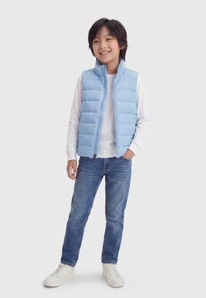 Gutt smiler og har på seg en lyseblå vattert vest, hvit langermet skjorte, blå jeans og hvite joggesko, står foran en ensfarget bakgrunn.