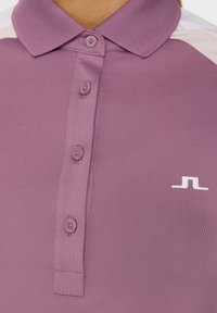 Polo shirt viola con colletto, dotato di una patta con due bottoni, texture liscia e un piccolo logo bianco sul petto.
