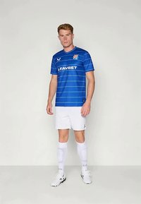 Mėlynas futbolo marškinėlis su horizontaliais dryžiais, kuriame yra logotipas ir rėmėjo tekstas, derinamas su baltais šortais ir kojinėmis, eksponuojamas ant vyriško modelio.