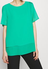 Blusa verde a maniche corte con un design a strati, realizzata in tessuto leggero. Presenta un colletto rotondo e una texture liscia.