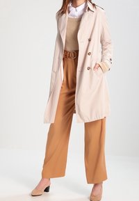 Beżowy trencz, dwurzędowy fason, z naszywkami na ramionach. Nosi się go na brązowym swetrze i szerokich brązowych spodniach. Gładka tekstura.