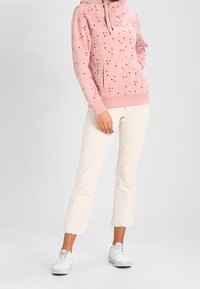 Felpa rosa con un motivo a macchie, dotata di cappuccio con cordino e tasca frontale, abbinata a pantaloni crema a zampa d'elefante e sneakers bianche.