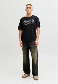 Sort t-shirt med "JACK & JONES" i mønstrede bogstaver, kombineret med falmede sorte jeans og beige mokkasin-sko. Modellen står foran en lys baggrund.