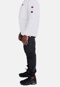 Man med en vit puffjacka med röd, vit och blå rosdesign på ärmarna, svarta cargobyxor och svarta sneakers.