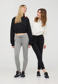 Due donne indossano felpe corte, una nera e una bianca, abbinate a pantaloni jogger grigi e neri. Entrambe indossano sneakers con dettagli in rete.