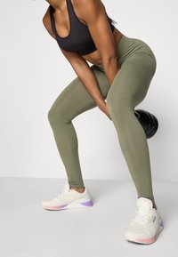 Sujeción deportiva negra, leggings verde oliva y zapatillas deportivas blancas con acentos morados. Incluye una pesa rusa en un fondo neutro.