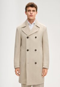 Beiger doppelt geknöpfter Mantel mit Fischgrätmuster, ausgestattet mit großen braunen Knöpfen und Seitentaschen, kombiniert mit einem weißen Hemd.