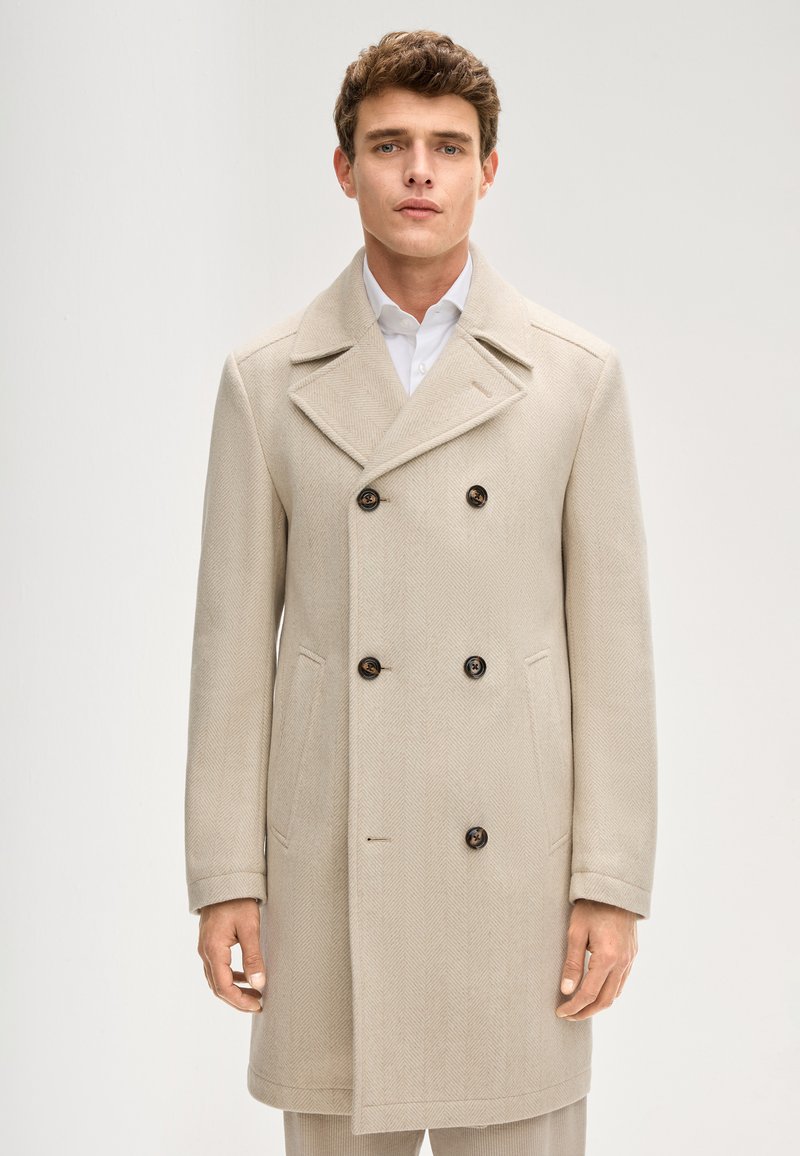 Beiger doppelt geknöpfter Mantel mit Fischgrätmuster, ausgestattet mit großen braunen Knöpfen und Seitentaschen, kombiniert mit einem weißen Hemd.