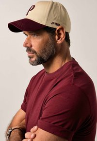 Cap with a cream crown and maroon brim, featuring a prominent "P" in maroon. Made of fabric, with adjustable strap at the back.
