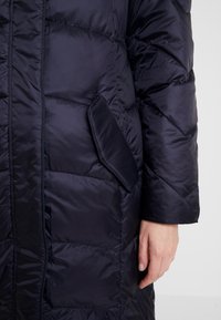 Námořnicky modrý péřový kabát s hladkým povrchem, vybavený boční kapsou a prošívaným designem. Rukáv má rovný střih a je mírně volnější.