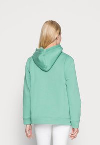 Mujer con cabello largo y rubio, vistiendo una sudadera verde menta sencilla y pantalones blancos, de pie de espaldas contra un fondo blanco.