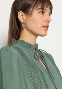 Blusa verde com um colarinho franzido, detalhe de laço e apliques de renda na parte frontal. Leve, com uma textura suave e um ajuste descontraído.