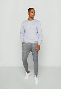 Homem de pé contra um fundo simples, vestindo um sweatshirt cinza claro, calças slim-fit com padrão xadrez e sapatilhas brancas, olhando para o lado.