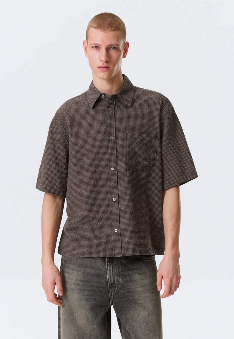 Jeune homme portant une chemise marron à manches courtes, boutonnée et texturée, ainsi qu'un jean gris délavé, se tenant contre un fond clair uni.