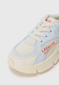 LACOSTE-sneakers har en lyseblå syntetisk overdel med mesh-detaljer, kremfargede snørebånd og en teksturert hvit såle. Merkevarens røde logo er en fremtredende detalj.