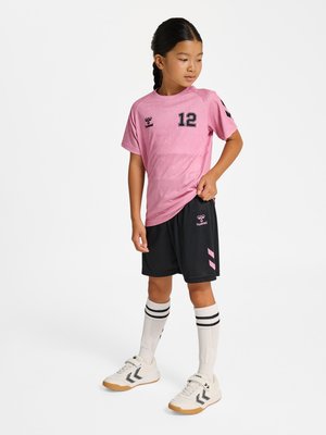 Joven niña con camiseta deportiva rosa número 12, pantalones cortos negros, calcetines blancos hasta la rodilla y zapatillas blancas, de pie con una mano en la cadera.