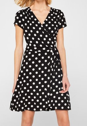 Schwarzes Wickelkleid mit weißen Punkten, kurzen Ärmeln, V-Ausschnitt und Taillenknoten, getragen von einer stehenden Person.