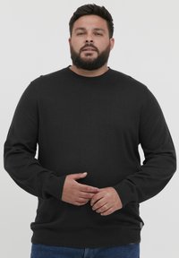 Pull en maille noir avec un col rond et des poignets côtelés. Le tissu semble doux et extensible, avec un léger éclat.