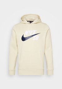 Beige Nike SB-huvtröja med framficka, justerbar dragsko på huvan och stor svart swoosh-logotyp med vit text på bröstet.