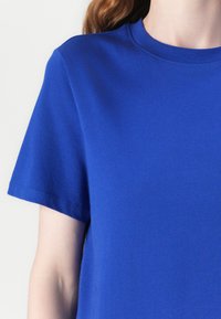 Sininen puuvillainen t-paita, jossa on pyöreä pääntie ja lyhyet hihat. Kankaan pinta näyttää sileältä, ja siinä on vain vähän näkyviä saumoja ilman kuvioita.