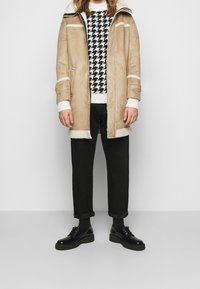 Abrigo de ante beige con terminaciones de borreguillo blanco, suéter de pata de gallo en blanco y negro, pantalones negros y zapatos negros de cordones.
