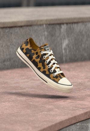 Converse sneaker met luipaardprint, witte veters en rubberen zool die boven een roze betonnen oppervlak zweeft.