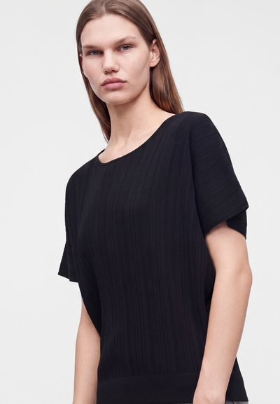 Jeune femme aux cheveux bruns raides portant un haut noir à manches courtes avec un subtil motif côtelé vertical sur un fond uni.