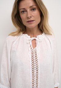 Bijela lanena bluza s okruglim izrezom, vezanjem sprijeda i dekorativnim čipkastim detaljima. Ima labave, napuhane rukave i teksturiranu tkaninu.