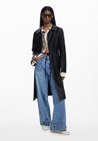 Cappotto trench in pelle nera con vita stretta, abbinato a pantaloni in denim blu oversize. Include una camicia a fantasia sotto e sneakers bianche.
