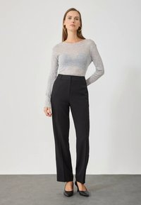 Donna che indossa un top trasparente grigio a maniche lunghe, pantaloni dritti neri a vita alta, tacchi neri e orecchini a cerchio dorati, in piedi contro un muro neutro.