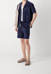 Uomo che indossa una camicia a maniche corte navy con motivo floreale e pantaloni coordinati, una canotta bianca, sandali beige e porta un tote bag beige.